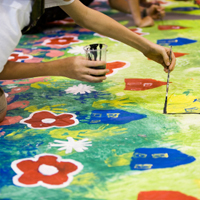 Ein großes Bild mit Häusern und Blumen wird von mehreren Kinderhänden mit Tusche gemalt.